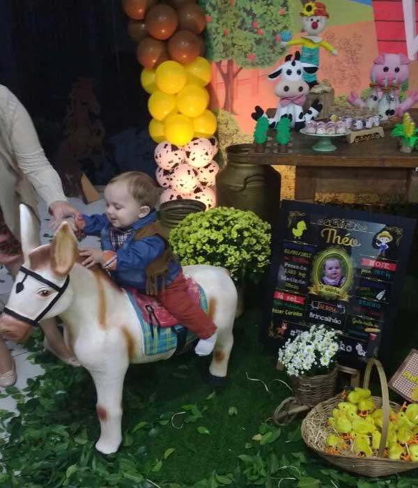 Aniversariante Théo vestido de cowboy com roupa xadrez, colete marrom de franja e calça jeans vermelha brincando de montar em cima do cavalinho de brinquedo