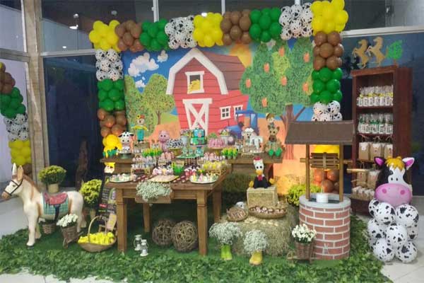 Decoração da festa infantil fazendinha com muitos balões coloridos, graminhas no chao e bonecos de pelúcia de animais e no canto direito um mini poço de água