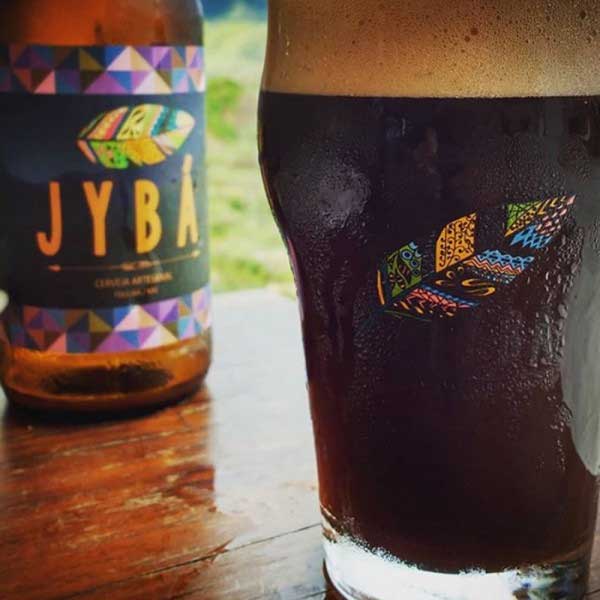 Copo com emblema da marca Jybá cheio de cerveja preta Stout e ao fundo uma garrafa dessa que está entre as melhores bebidas de inverno