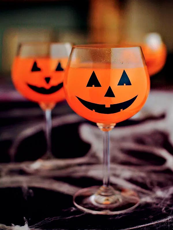 Taça transparente cheia de refrigerante de laranja e por fora colado olhos e boca de papel imitando uma abóbora de decoração de Halloween